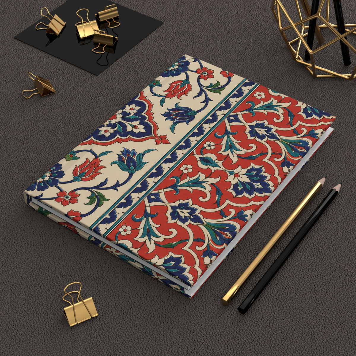 A notebook with a traditional red, blue, and green floral pattern on a cream background, styled on a dark gray textured surface with gold binder clips, pencils, and a geometric gold object.