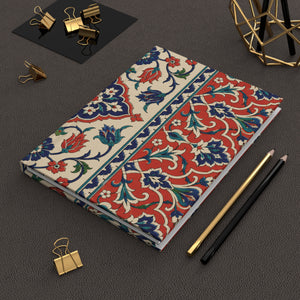 A notebook with a traditional red, blue, and green floral pattern on a cream background, styled on a dark gray textured surface with gold binder clips, pencils, and a geometric gold object.