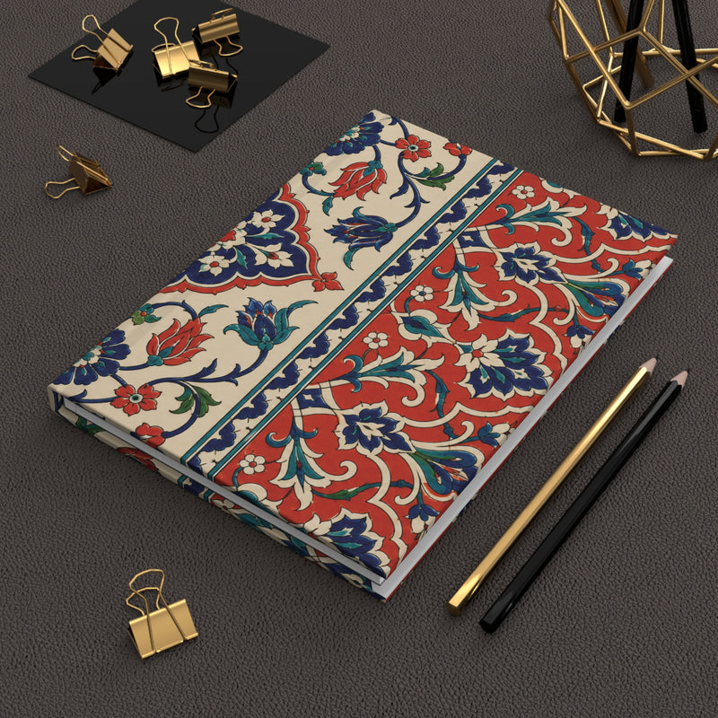 A notebook with a traditional red, blue, and green floral pattern on a cream background, styled on a dark gray textured surface with gold binder clips, pencils, and a geometric gold object.