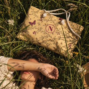 A beige tote bag with a floral print, butterfly motif, and white rope handles lies on a grassy lawn next to a person lying on the grass.