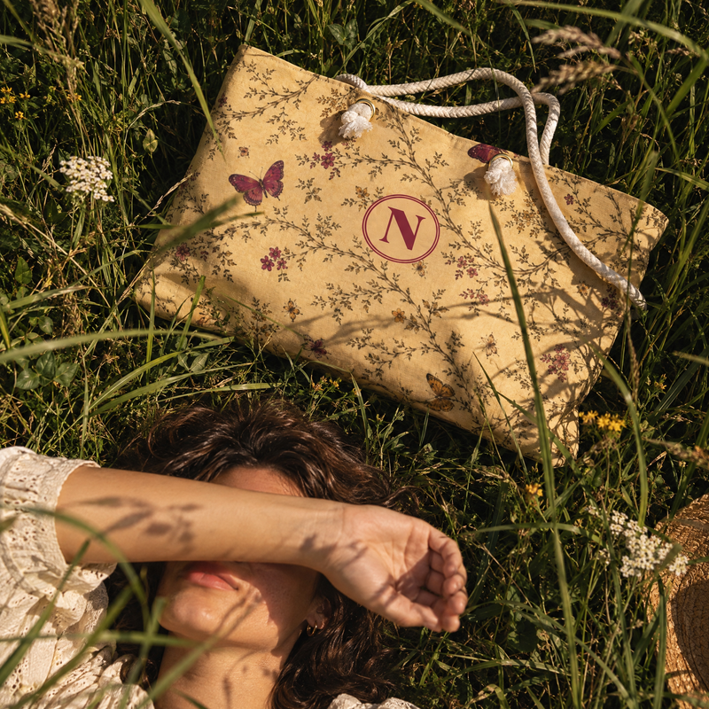 A beige tote bag with a floral print, butterfly motif, and white rope handles lies on a grassy lawn next to a person lying on the grass.
